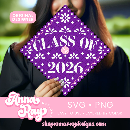 A person in a graduation gown holds a purple graduation cap decorated with white floral and geometric papel‑picado–style patterns and the text “Class of 2026” in bold white letters.